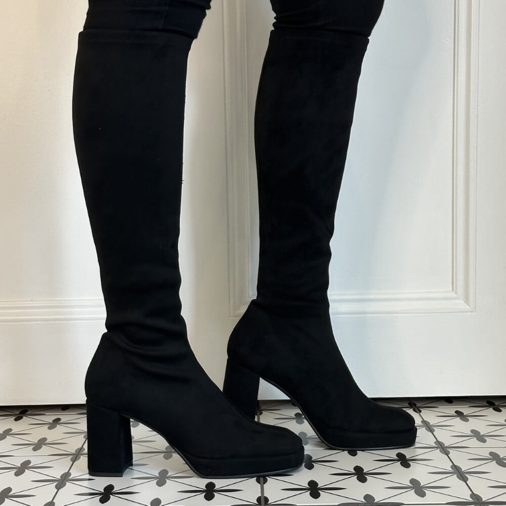 A pair of Miss Elastic black suede-like knee high boots with chunky block heels, shown worn with dark pants against a white panelled wall and patterned tile floor.