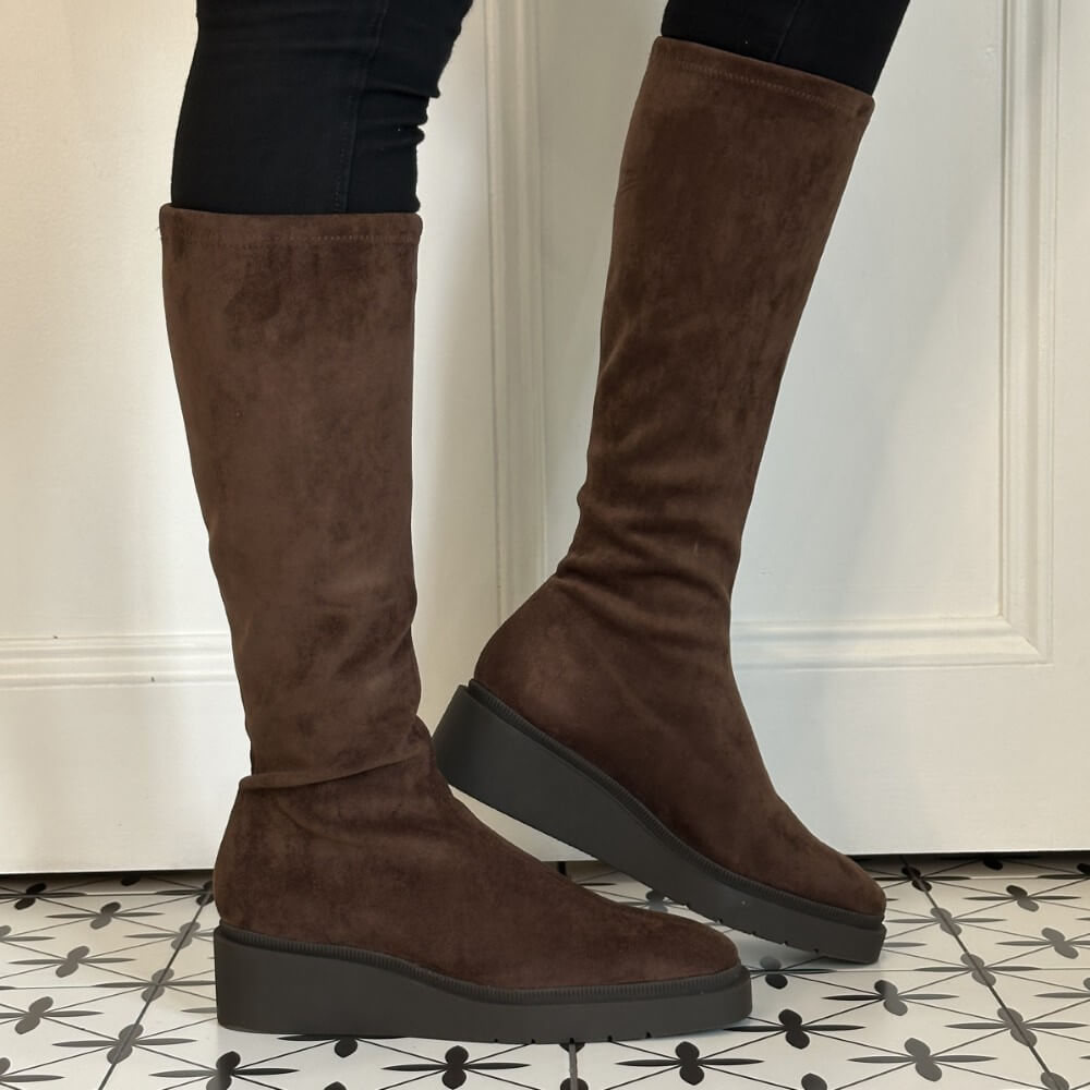 A pair of  Miss Elastic knee-high brown suede boots with chunky black platform soles, shown on a model wearing black leggings in front of a white panelled door.