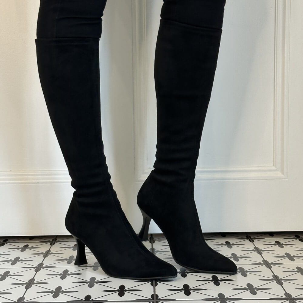 Miss Elastic Black knee high heeled boots with a pointed toe and slim kitten heel, shown on a model wearing black leggings against a white panelled wall and patterned tile floor.