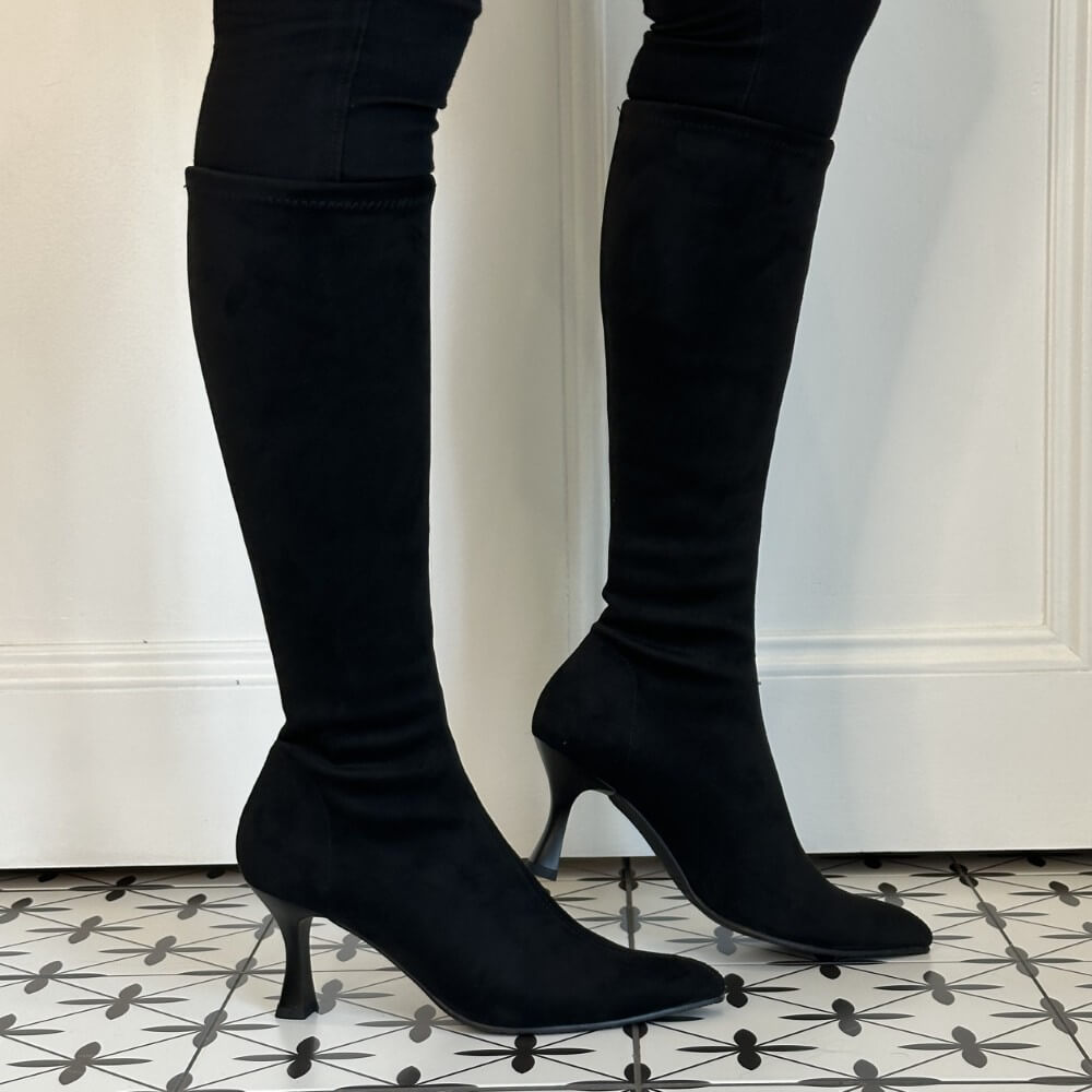 Miss Elastic Black knee high heeled boots with a pointed toe and slim kitten heel, shown on a model wearing black leggings against a white panelled wall and patterned tile floor.