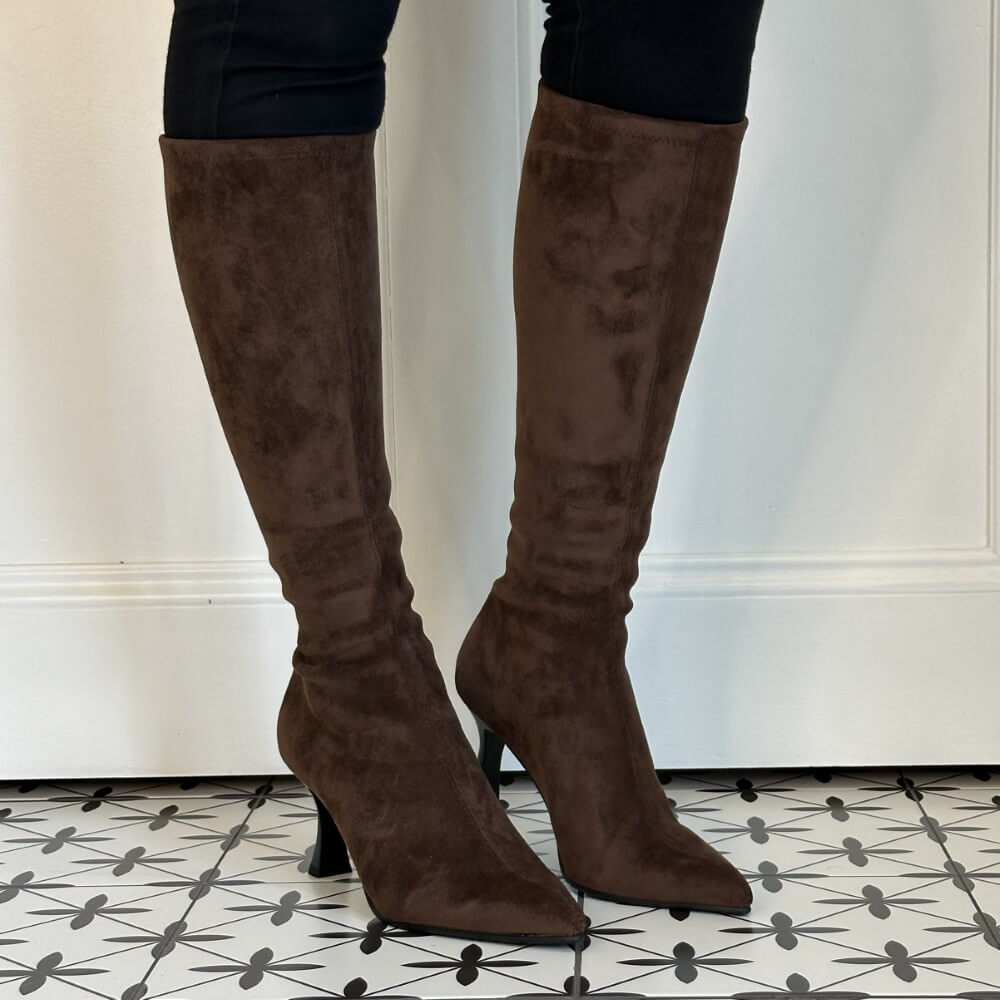 A pair of Miss Elastic dark brown suede knee-high heeled boots worn with black pants, standing on a black and white patterned tile floor in front of a white wall.