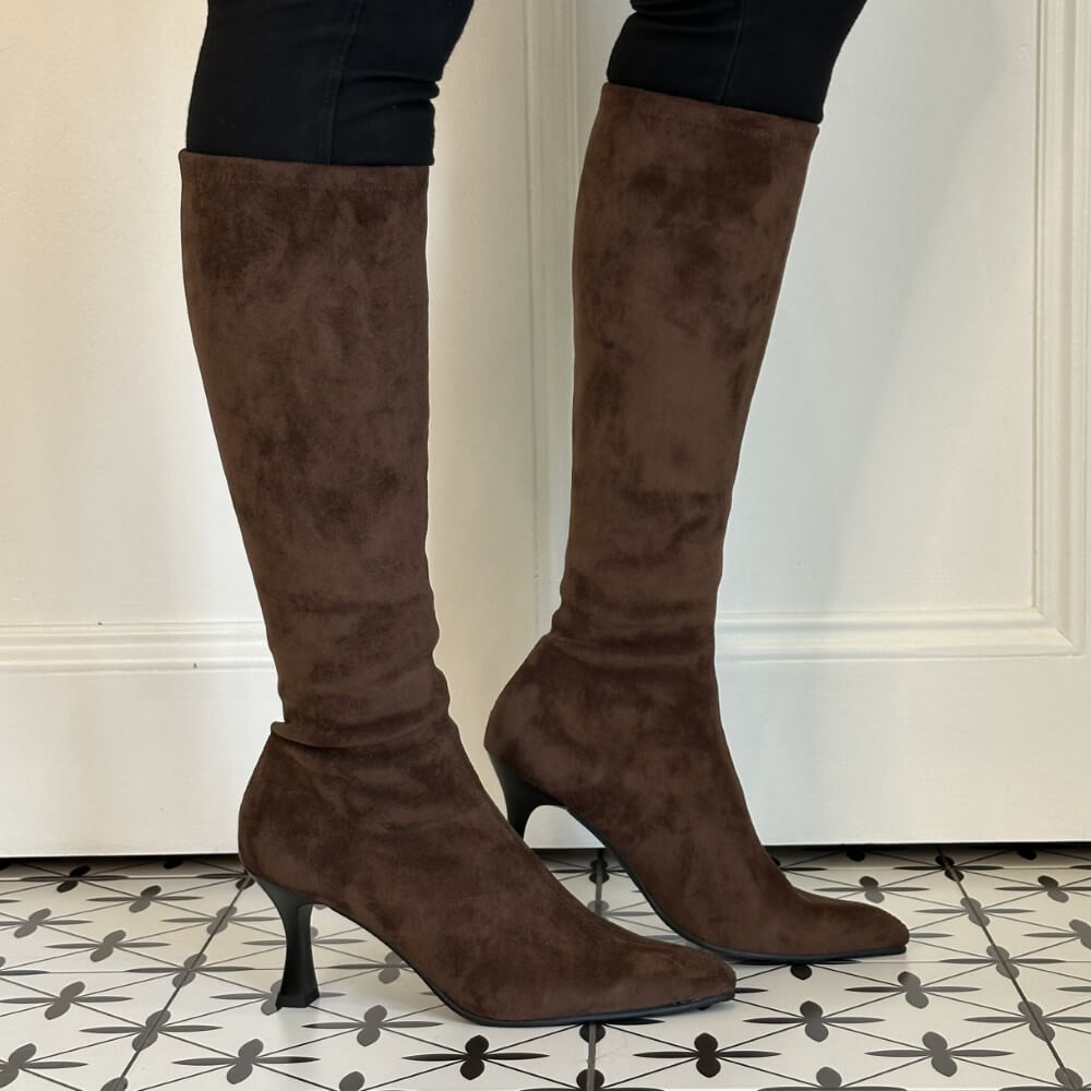 A pair of Miss Elastic knee-high brown suede boots with a kitten heel, worn with black leggings, standing on a black and white patterned tile floor in front of a white panelled wall.