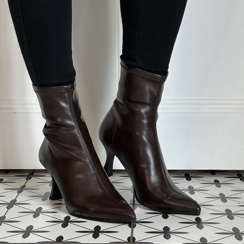 A pair of Miss Elastic dark brown stretch ankle boots with pointed toes and slim high stiletto heels, shown on a model wearing black pants against a white wall and patterned tile floor.