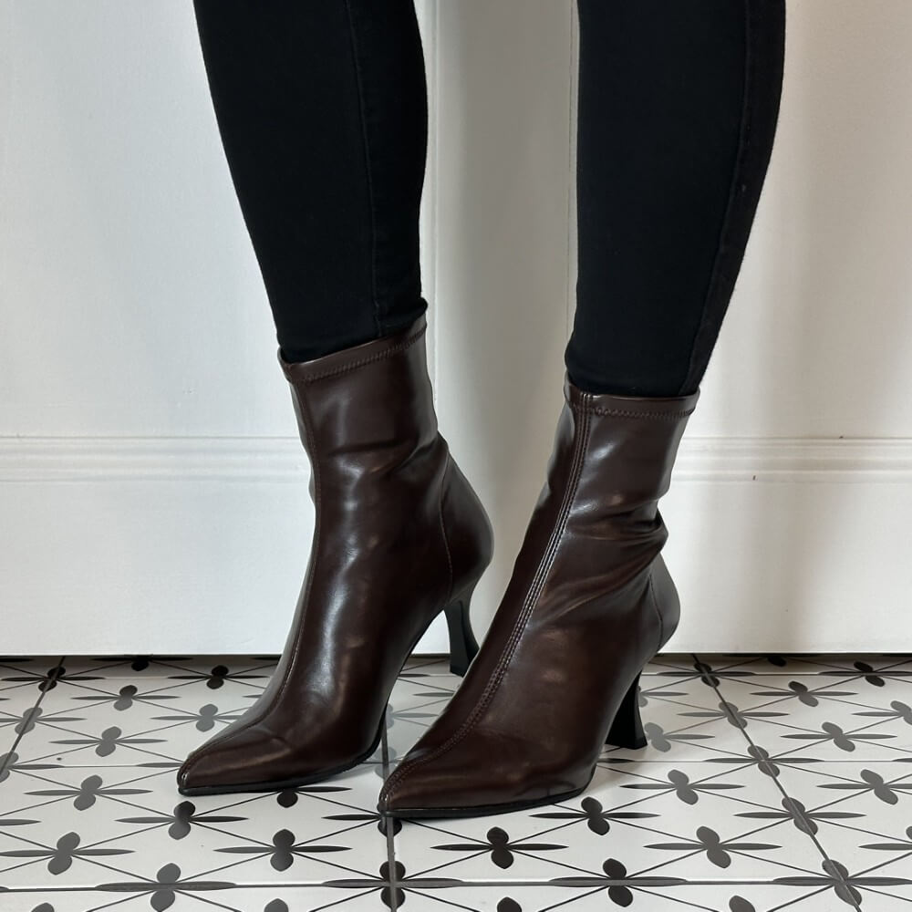 A pair of Miss Elastic dark brown stretch ankle boots with pointed toes and slim high stiletto heels, shown on a model wearing black pants against a white wall and patterned tile floor.