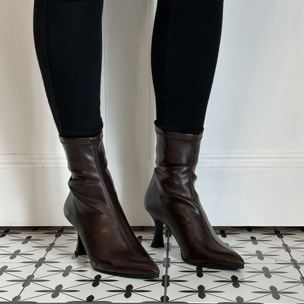 A pair of Miss Elastic dark brown stretch ankle boots with pointed toes and slim high stiletto heels, shown on a model wearing black pants against a white wall and patterned tile floor.