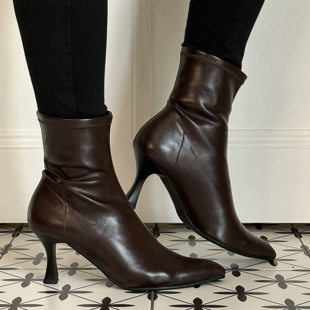 A pair of Miss Elastic dark brown stretch ankle boots with pointed toes and slim high stiletto heels, shown on a model wearing black pants against a white wall and patterned tile floor.