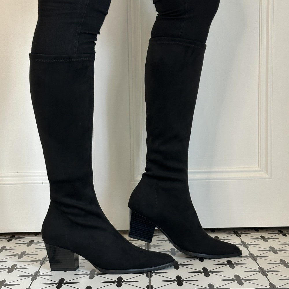  Miss Elastic Black suede knee high boots with a block heel, shown on a model wearing black skinny jeans against a white panelled wall and black-and-white patterned tile floor.