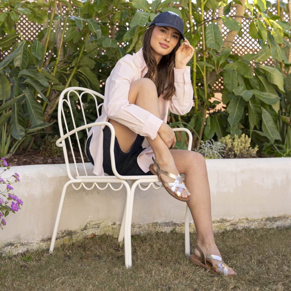 Woman wearing Oh My Sandals gold sandals sitting on a white chair outdoors with greenery in the backgrounds 