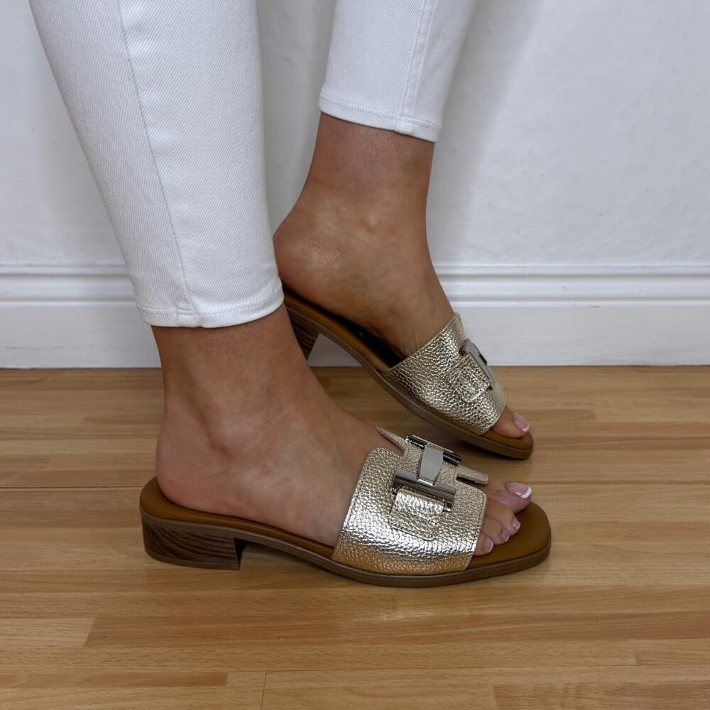 Oh My Sandals 5890 Gold sandals worn with white pants on a wooden floor.