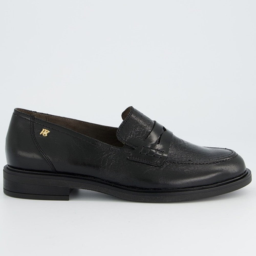 A Paul Green black leather penny loafer shoe with a low block heel and a small gold monogram logo on the heel counter, shown from a side profile against a light gray background.