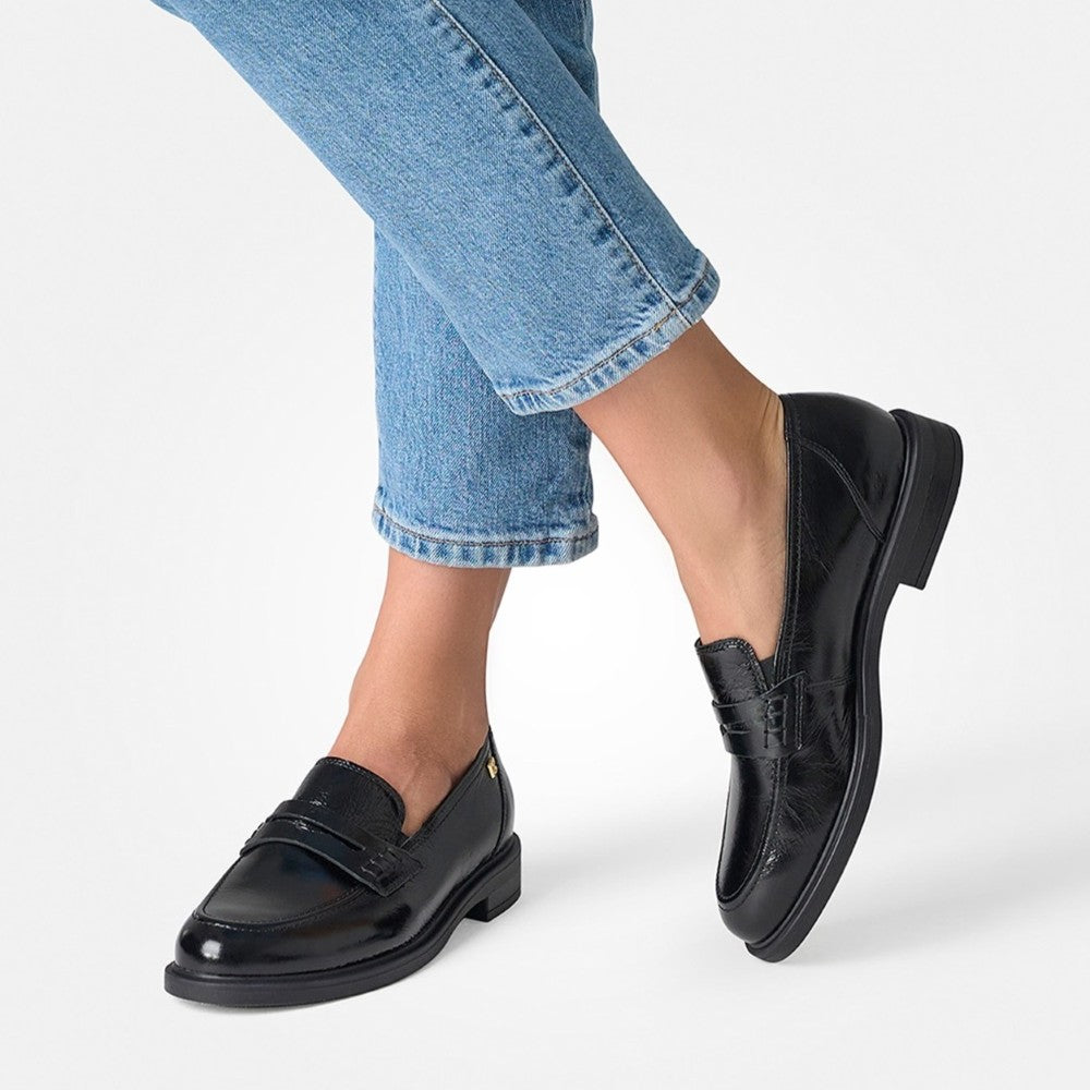A person wearing Paul Green black patent leather penny loafers with light blue cuffed jeans, shown from the knees down against a clean white studio background.