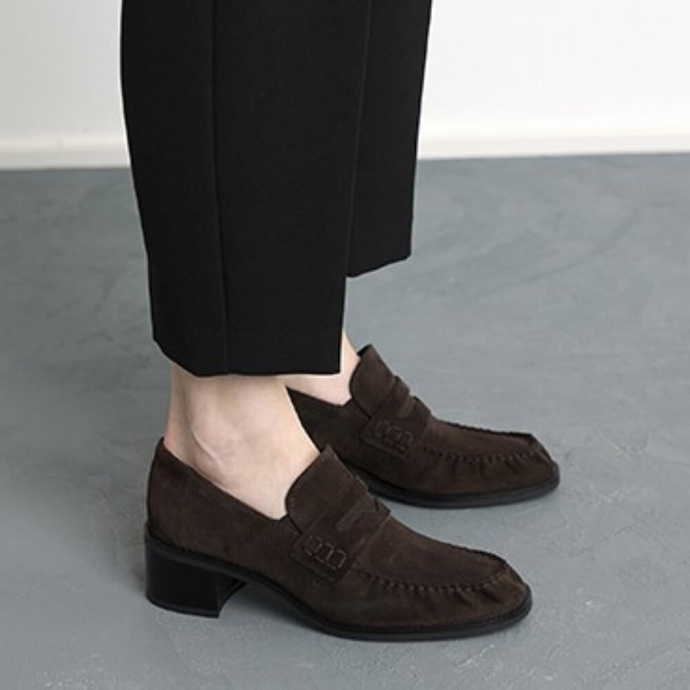 A person wearing Paul Green dark brown suede penny loafers with low block heels and black trousers, standing on a grey floor against a white wall.