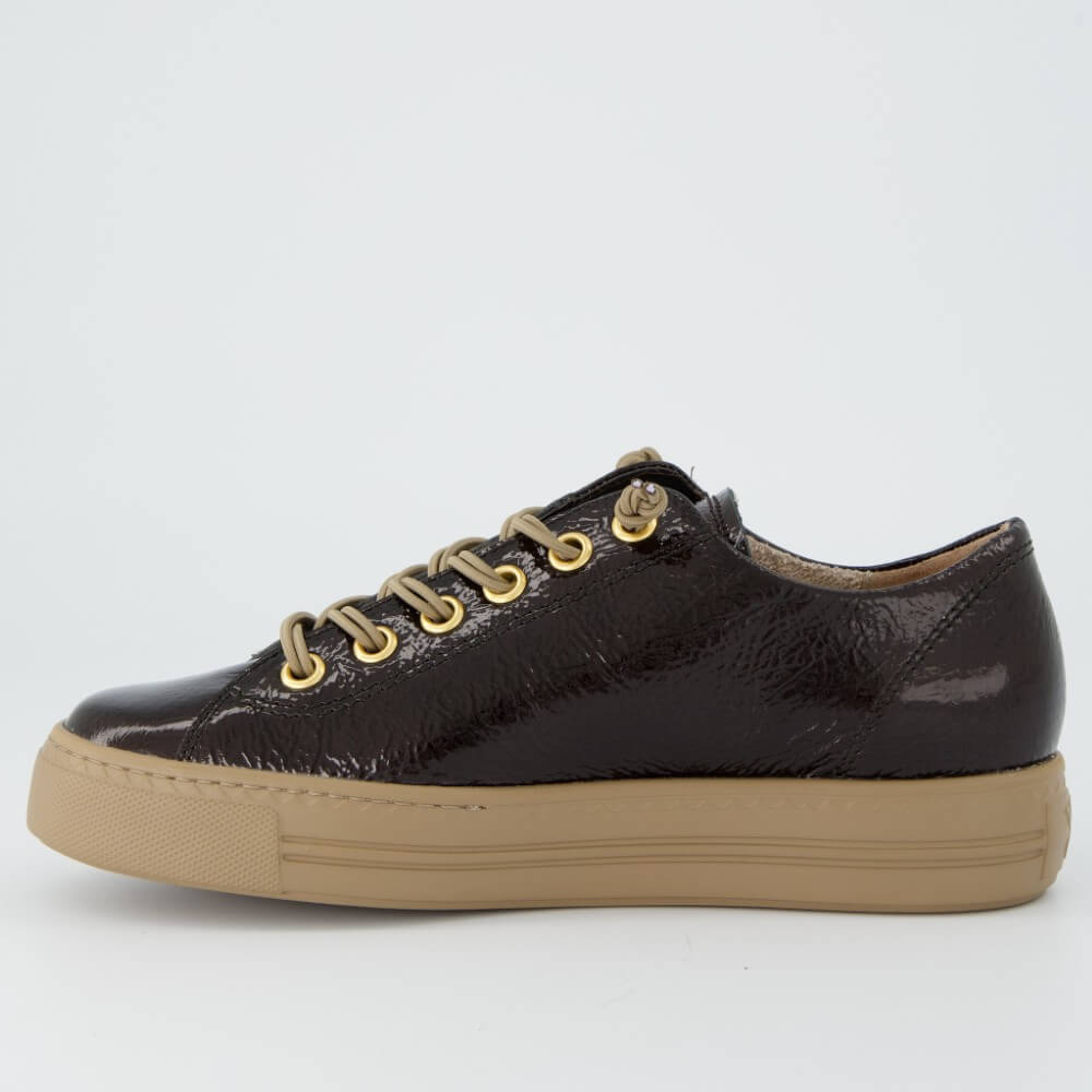 A Paul Green low-top sneaker in glossy dark brown textured leather with beige laces threaded through gold-tone eyelets and a thick beige rubber sole, shown on a light grey background.