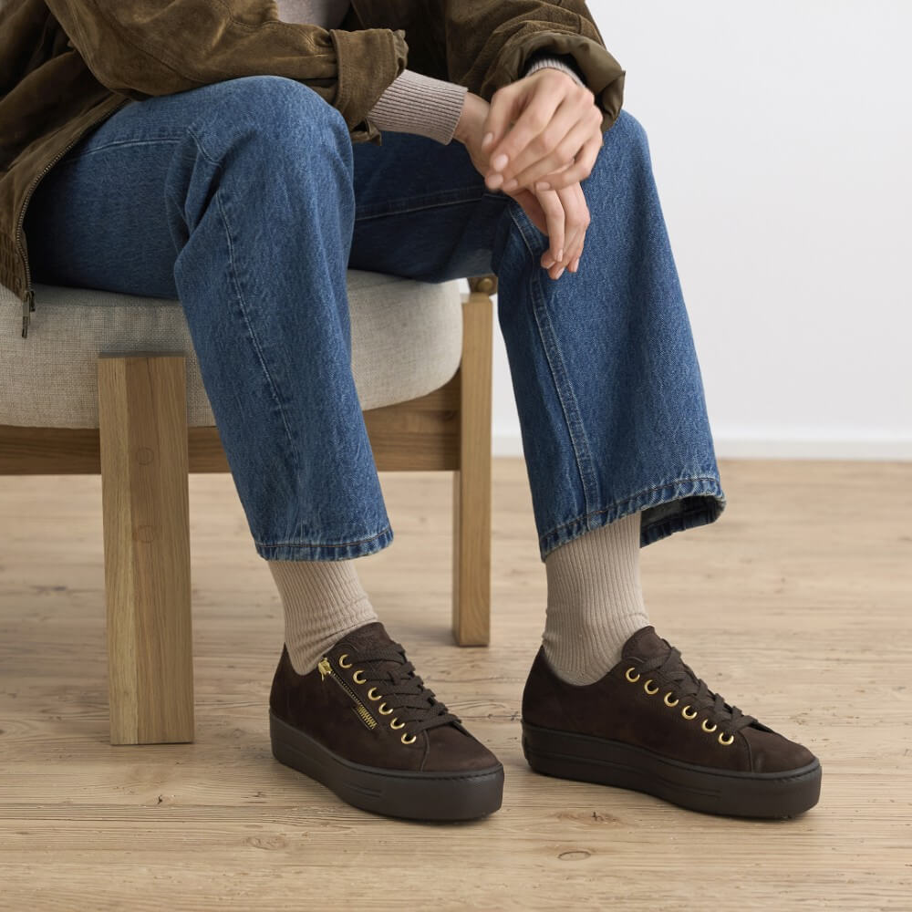 A person seated on a wooden chair wearing Paul Green dark brown suede platform sneakers with gold eyelets, blue jeans, and beige socks in a bright indoor setting with light wood flooring.