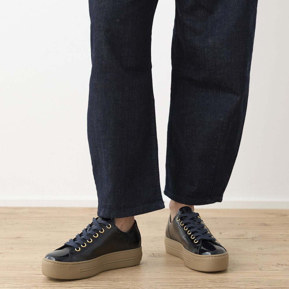 A person wearing dark navy wide-leg jeans and Paul Green glossy navy low-top platform sneakers with gold eyelets, standing on a light wood floor against a white wall.