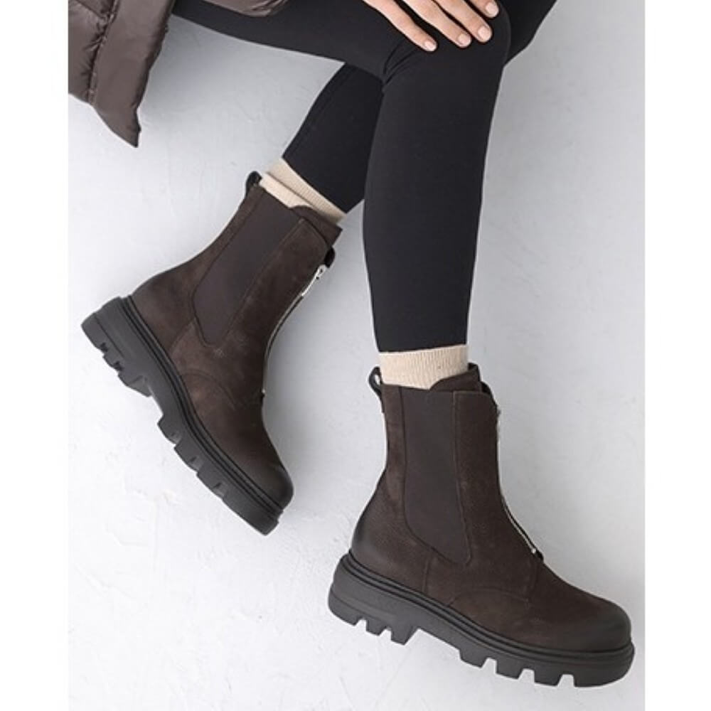 A person wearing Paul Green dark brown ankle boots with elastic side panels and chunky lug soles, paired with black leggings and beige socks, seated against a light grey background.