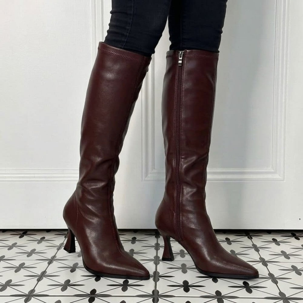 A pair of Pedro Anton Wine knee-high leather boots with pointed toes and kitten heels, shown on a model's legs against a white panelled door and patterned tile floor.