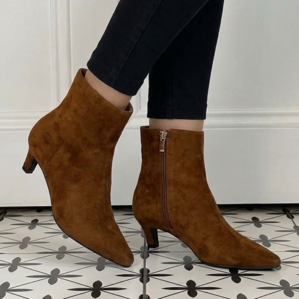 A pair of Pedro Anton Tan suede ankle boots with kitten heels and side zippers, shown on a model wearing black pants against a white panelled wall and patterned tile floor.