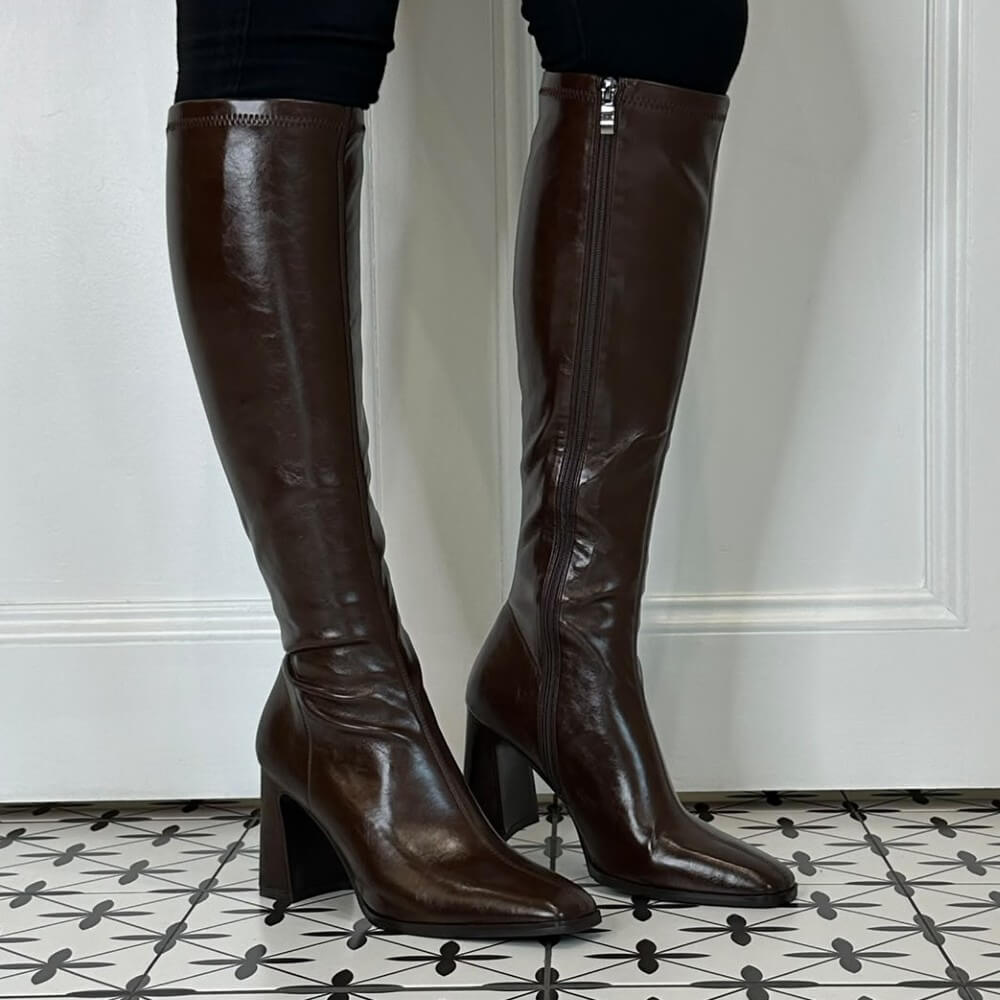 A pair of Pedro Anton dark brown knee-high leather boots with a side zipper and block heel, shown on a model's feet against a white panelled wall and patterned tile floor.