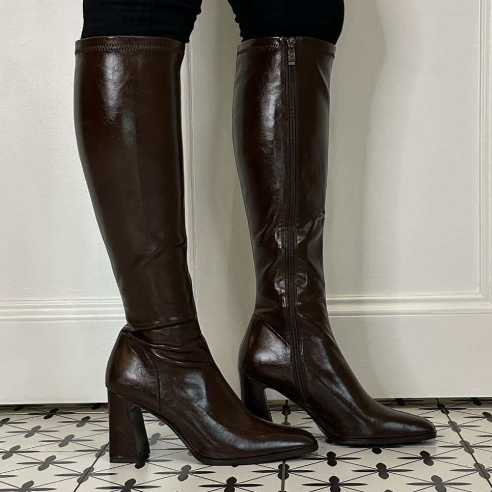 A pair of Pedro Anton dark brown knee-high leather boots with a side zipper and block heel, shown on a model's feet against a white panelled wall and patterned tile floor.