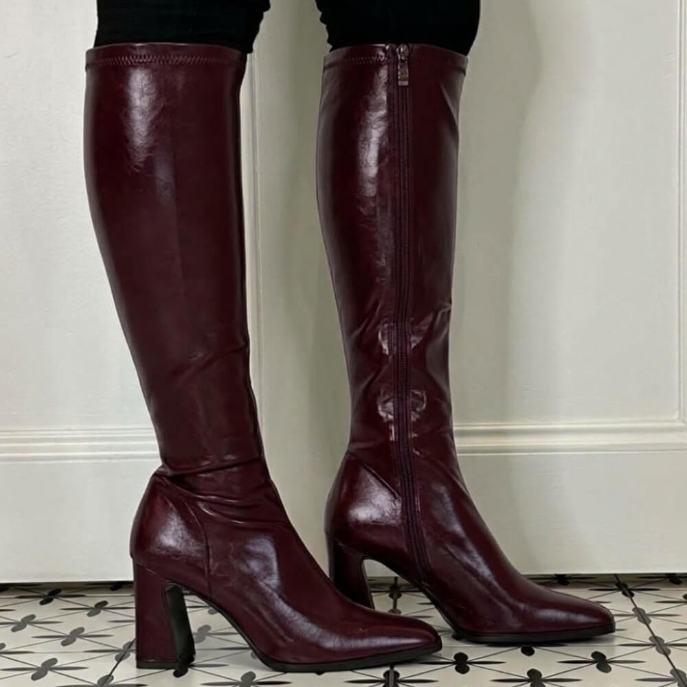 A pair of Pedro Anton wine knee-high leather boots with a side zipper and block heel, shown on a model's feet against a white panelled wall and patterned tile floor.
