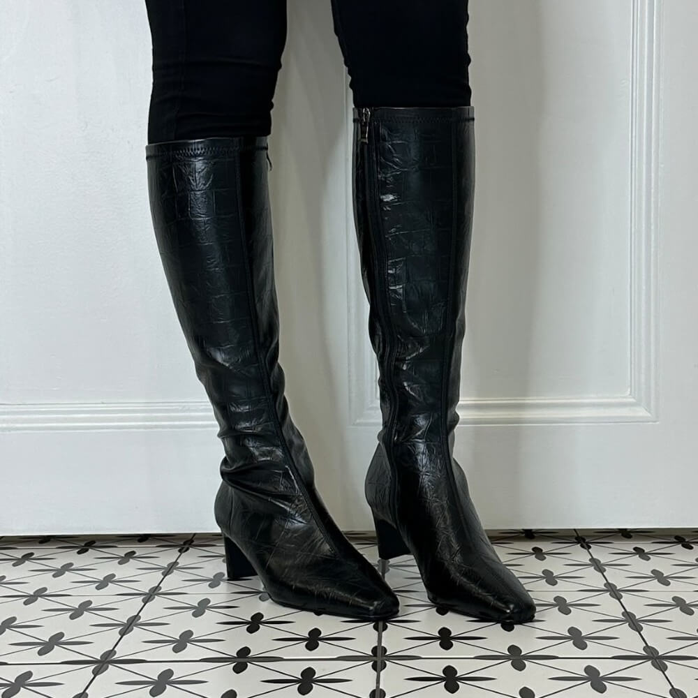 A person wearing Pedro Anton knee-high dark brown crocodile-embossed leather boots with kitten heels, standing on a black and white patterned tile floor in front of a white panelled wall.