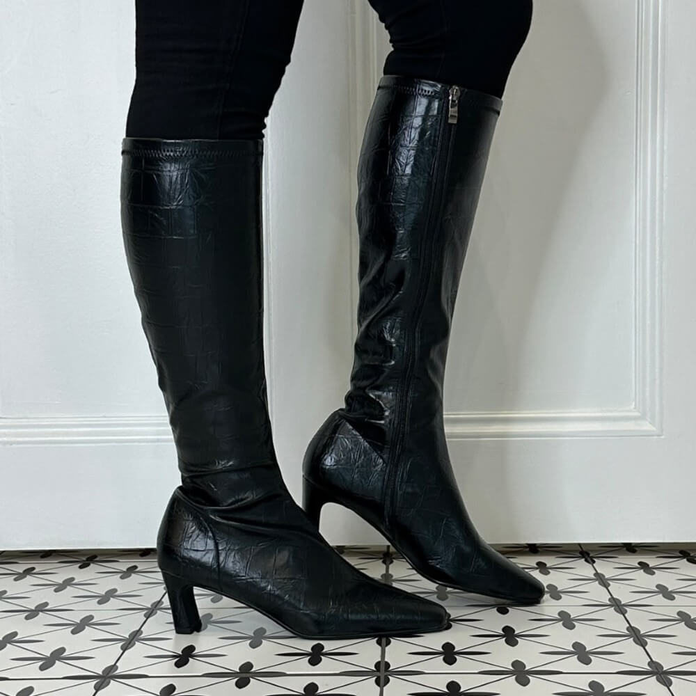 A pair of Pedro Anton black knee-high boots with a textured crocodile-embossed finish, shown on a model's legs against a white panelled wall and a black-and-white patterned tile floor.