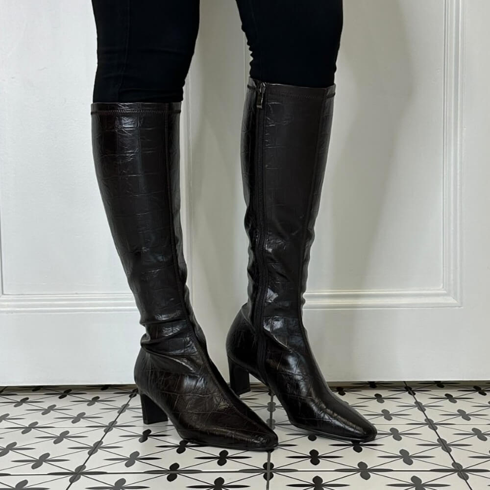 A person wearing Pedro Anton knee-high dark brown crocodile-embossed leather boots with kitten heels, standing on a black and white patterned tile floor in front of a white panelled wall.