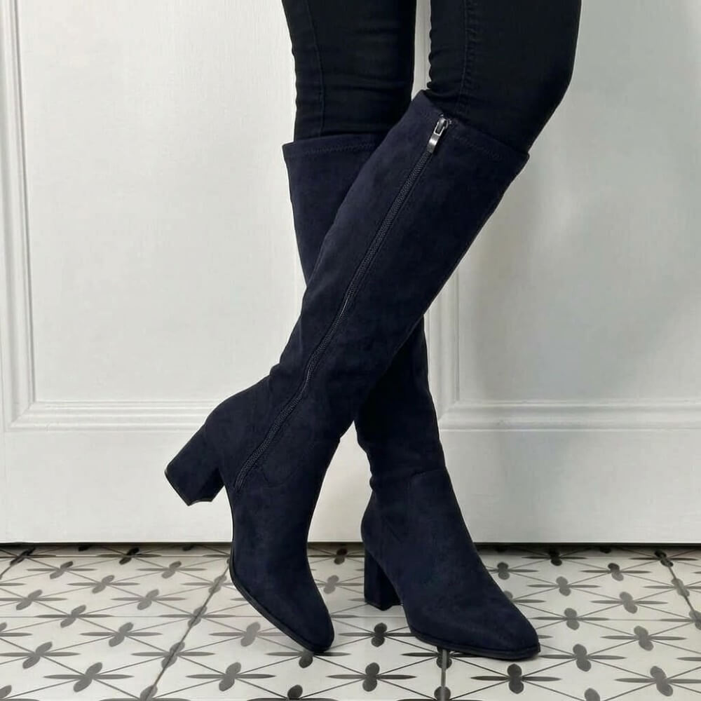 A pair of Pedro Anton dark navy blue suede knee-high boots with block heels, worn with black skinny jeans, standing on a black and white patterned tile floor in front of a white panelled wall.