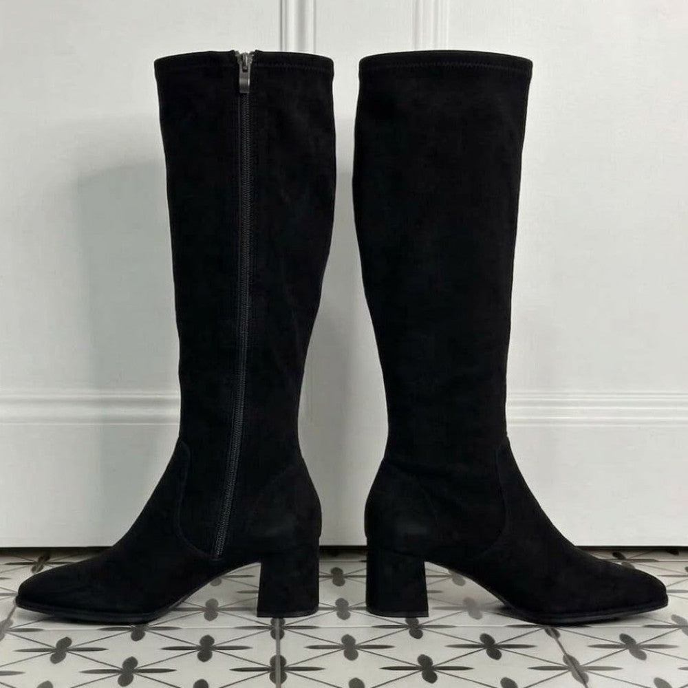 A pair of Pedro Anton black suede knee-high boots with block heels and side zippers, displayed on a patterned tile floor against a white panelled wall.