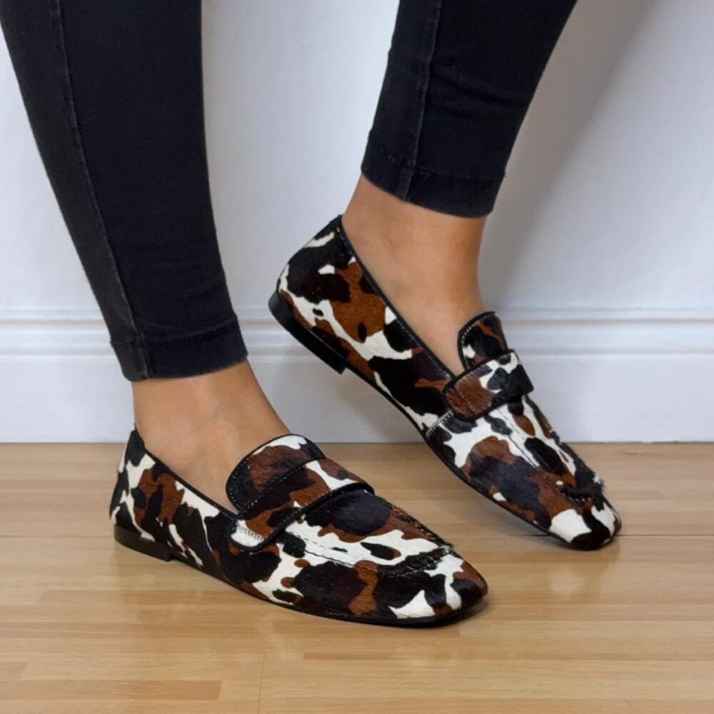 Cow print loafers worn with black pants on a wooden floor. Pedro Anton Bari Cow Print Loafers