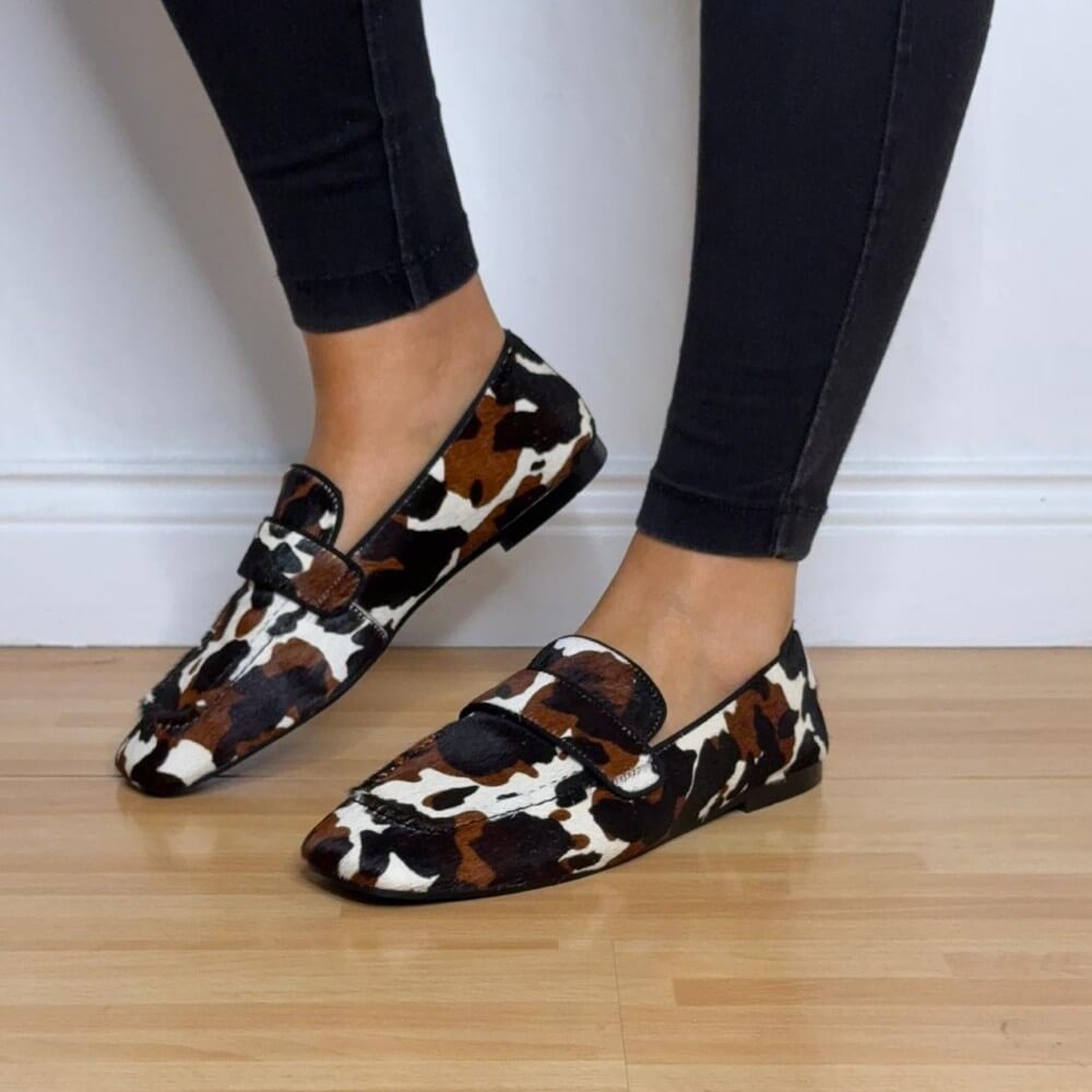 Cow print loafers worn with black pants on a wooden floor. Pedro Anton Bari Cow Print Loafers