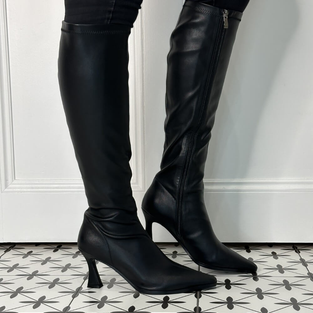 A pair of Pedro Anton black leather knee-high boots with pointed toes and kitten heels, shown worn with black pants against a white panelled wall and patterned tile floor.