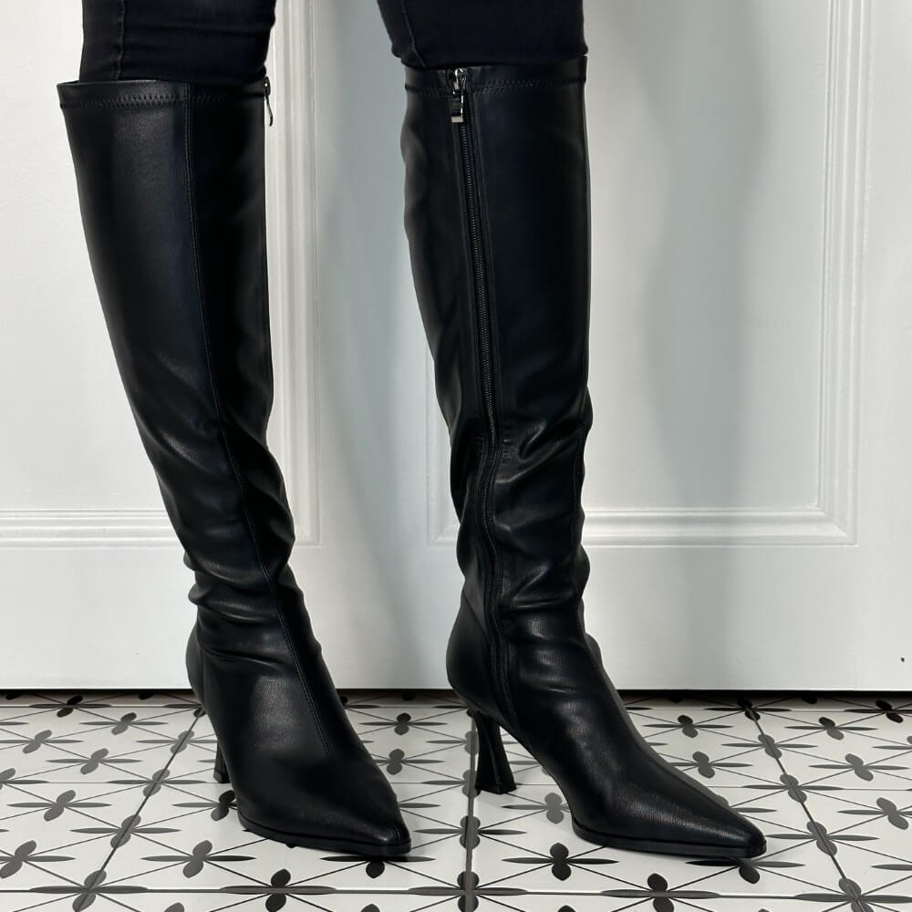 A pair of Pedro Anton black leather knee-high boots with pointed toes and kitten heels, shown worn with black pants against a white panelled wall and patterned tile floor.
