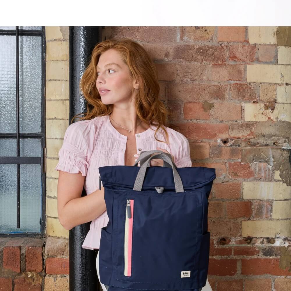 Woman holding a Roka navy blue backpack bag against a brick wall