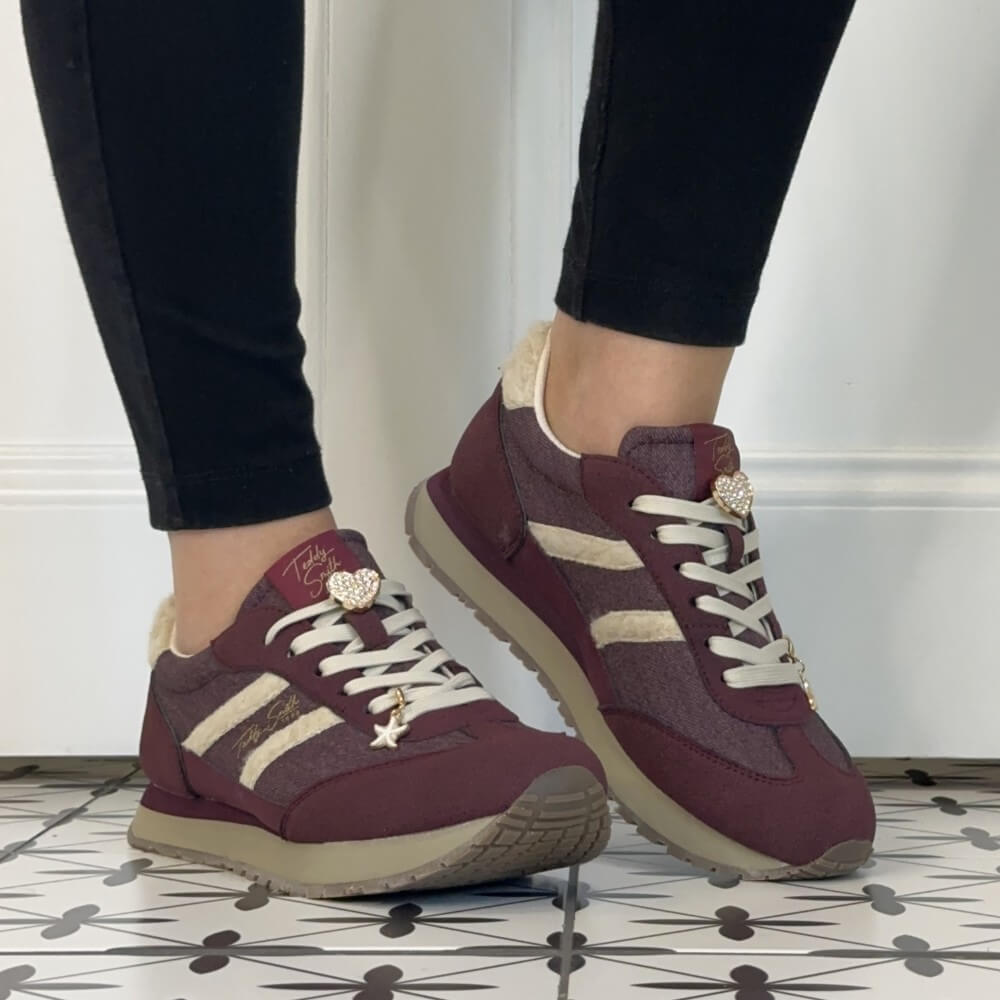 A person wearing Teddy Smith burgundy and beige low-top sneakers with black pants, standing on a patterned tile floor against a white wall.