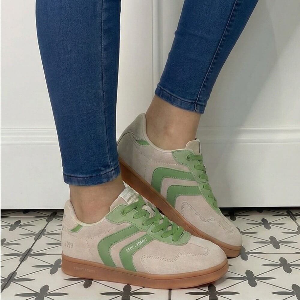 Beige sneakers with green accents worn with blue jeans on a tiled floor.
