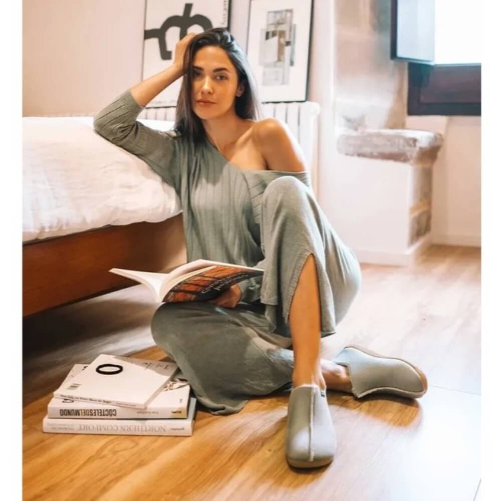 A woman sits on a wooden floor in a bedroom setting, wearing a sage green one-shoulder long-sleeve dress with matching wide-leg pants and slip-on Toni Pons Slippers. She is holding an open magazine, with a stack of books on the floor beside her.