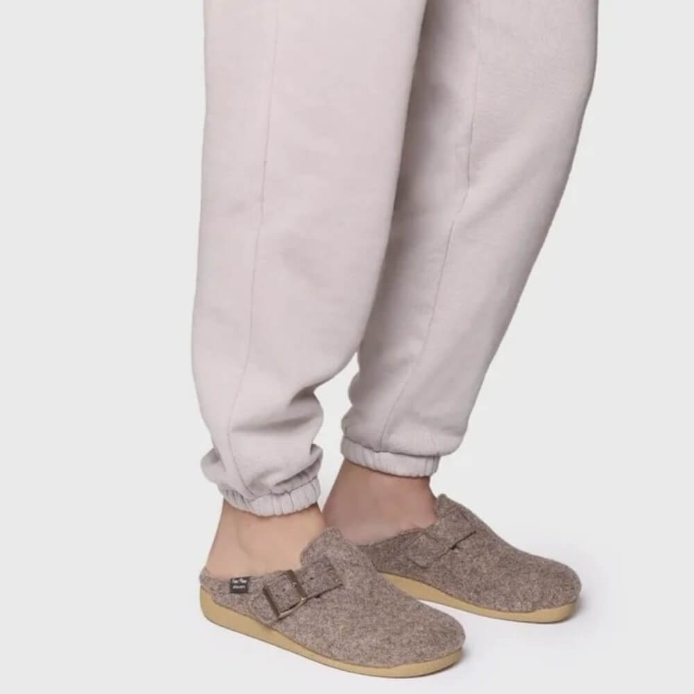 A person wearing light beige jogger pants and Toni Pons taupe-brown felt clog slippers with straps and tan soles, shown from the calves down against a plain light grey studio background.