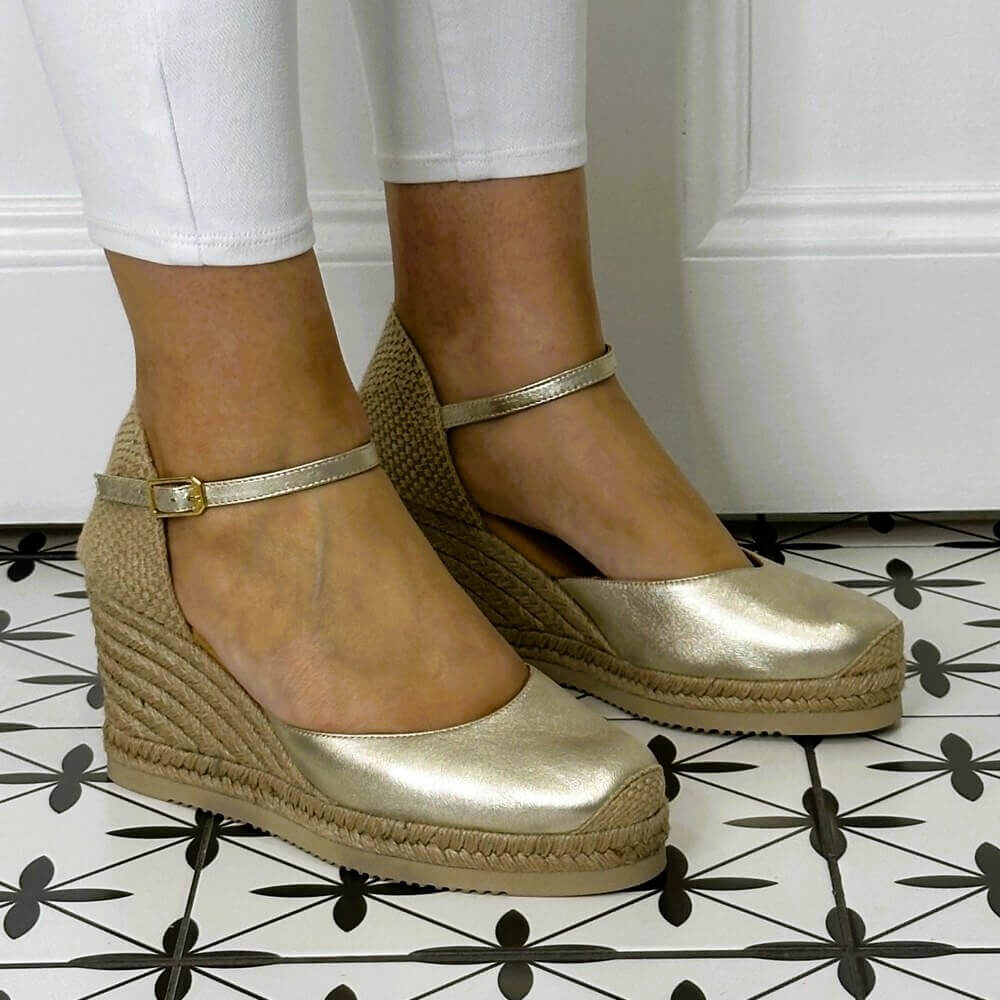 Gold wedge sandals with jute sole worn by a person on a patterned floor.