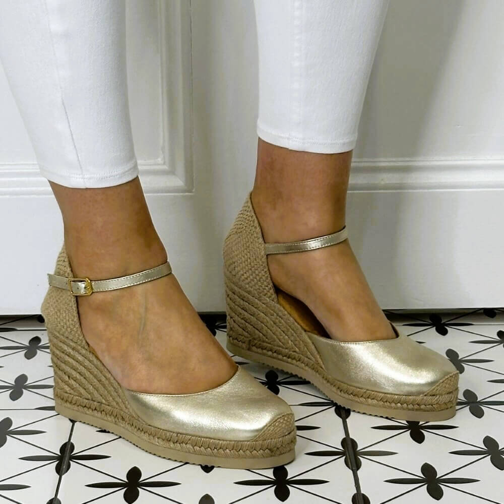 Gold wedge sandals with jute sole worn by a person on a patterned floor.