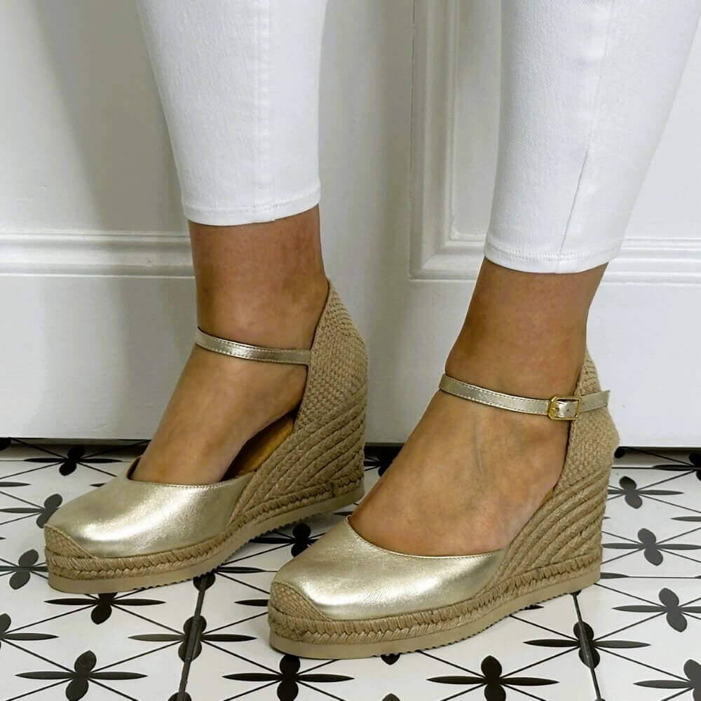 Gold wedge sandals worn with white pants on a patterned floor.