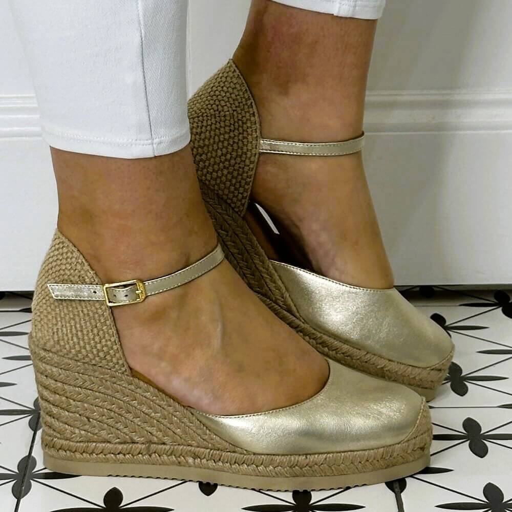 Gold wedge sandals with jute sole worn by a person on a patterned floor.