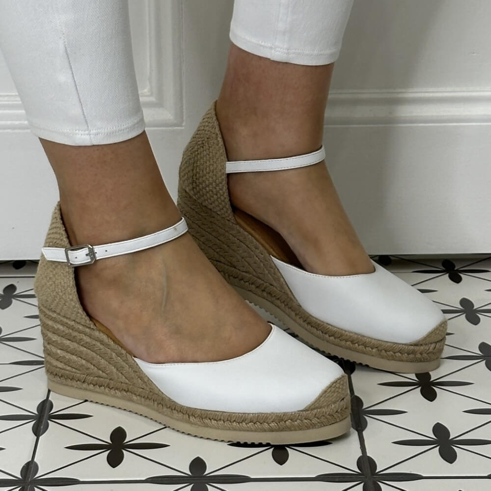 Unisa Caceres White wedge sandals with brown soles worn by a person on a tiled floor.