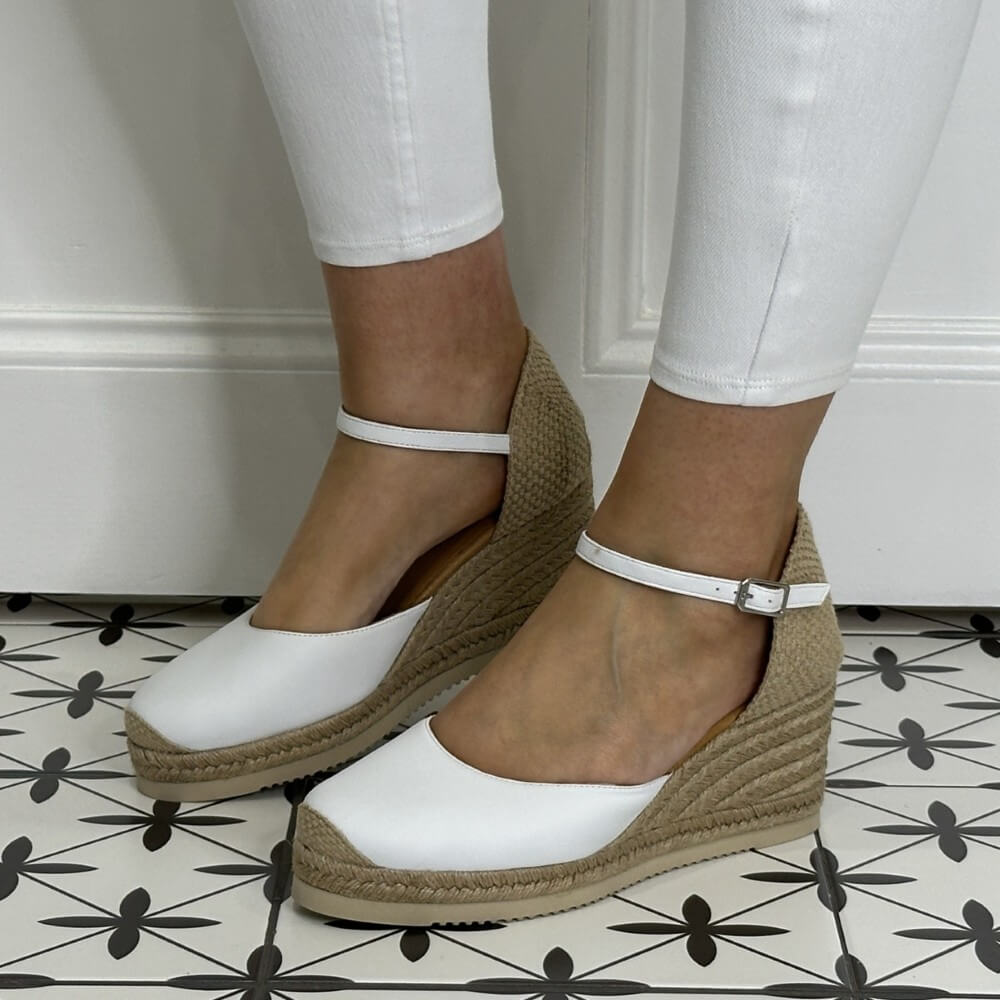 Unisa Caceres White wedge sandals with brown soles worn by a person on a tiled floor.