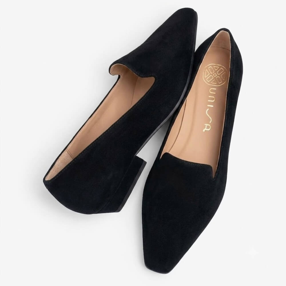 A pair of Unisa black suede pointed-toe flats with low block heels, displayed on a clean white background. One shoe is oriented upright, and the other lies flat on its side, showing the tan insole with the brand name 'UNISA' printed in gold.