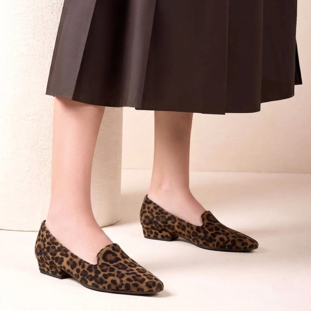 A person wearing Unisa leopard-print slip-on loafers with low block heels, paired with a dark brown pleated midi skirt, standing against a light beige textured wall.
