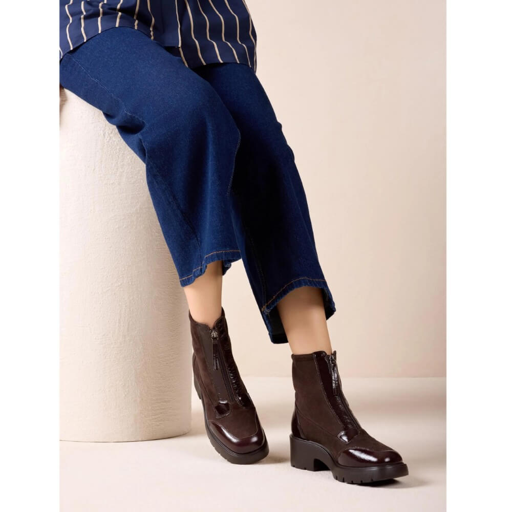 A person seated on a cylindrical stool wearing Unisa dark brown ankle boots with side zippers and dark blue cropped pants against a plain beige studio background.