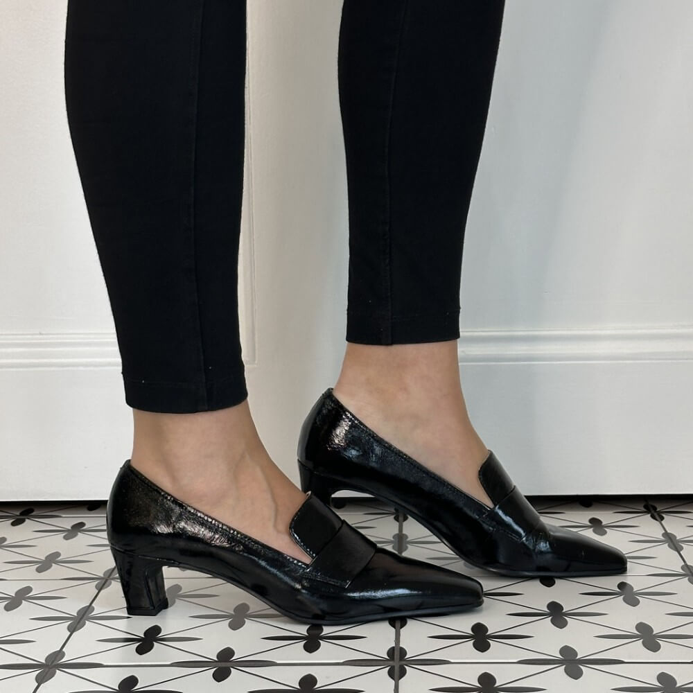 A person wearing Unisa black patent leather pointed-toe mule heels with a low block heel, paired with black leggings, standing on a black and white patterned tile floor against a white wall.