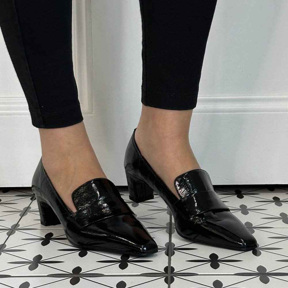 A person wearing Unisa black patent leather pointed-toe mule heels with a low block heel, paired with black leggings, standing on a black and white patterned tile floor against a white wall.